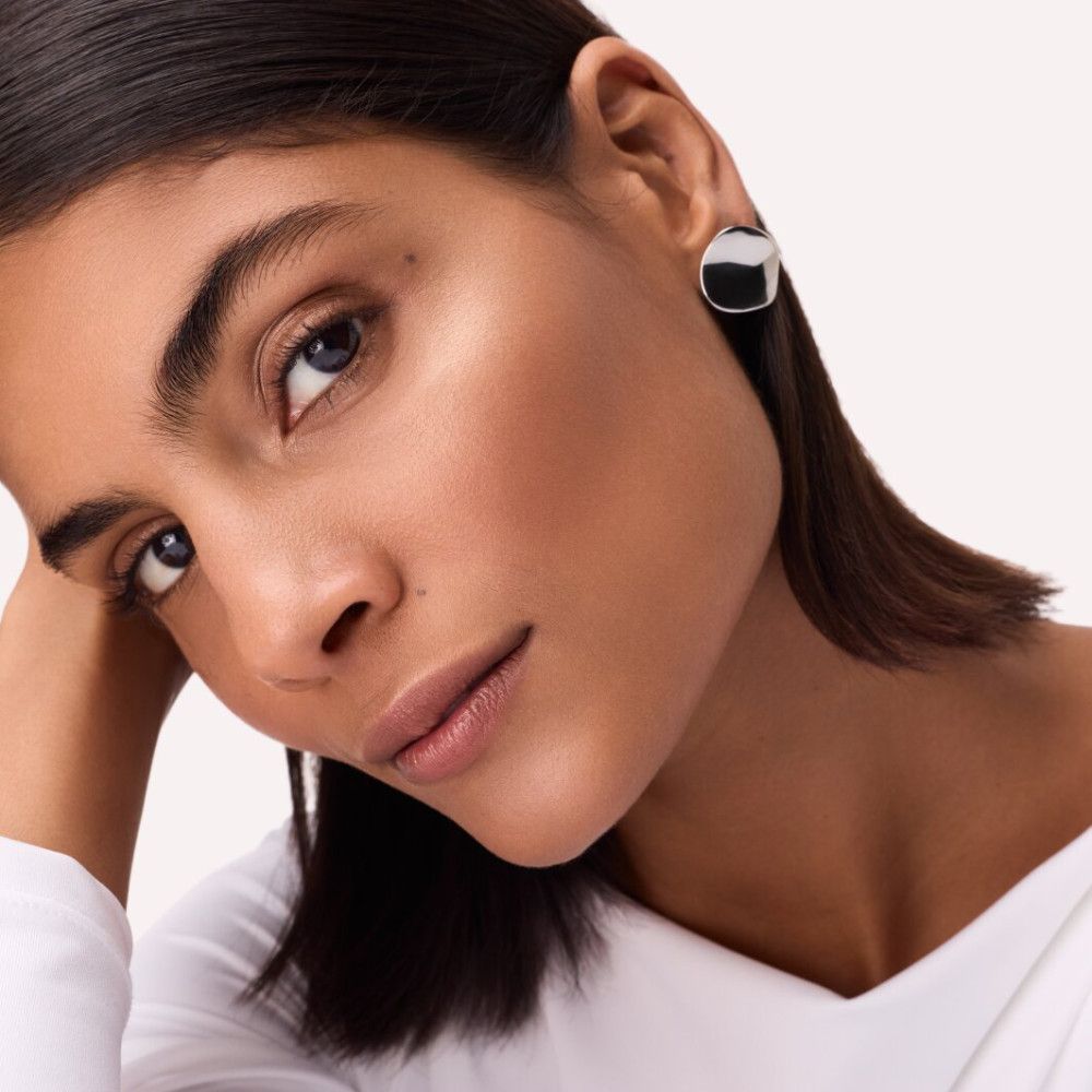 Porträt einer Frau mit Highlighter aufgetragen. Die Haut hat einen Glanz. Sie trägt runde Ohrringe und ein weißes Oberteil.