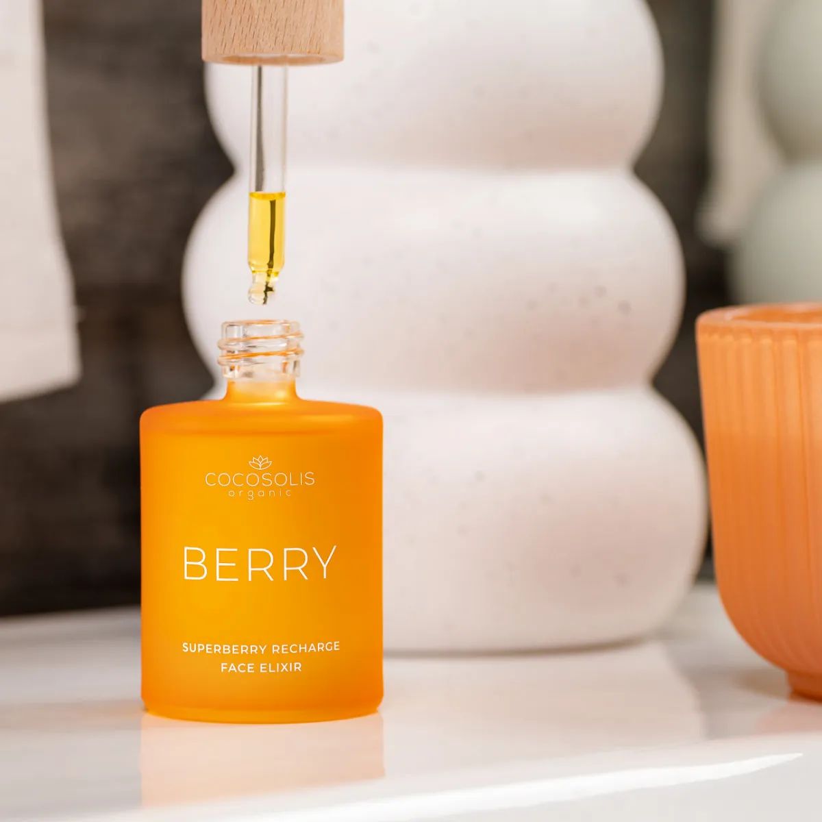 Orangefarbene Glasflasche mit Pipette, die auf einem Regal steht. Beschriftung: Cocosolis, Berry, Superberry Recharge Face Elixir.