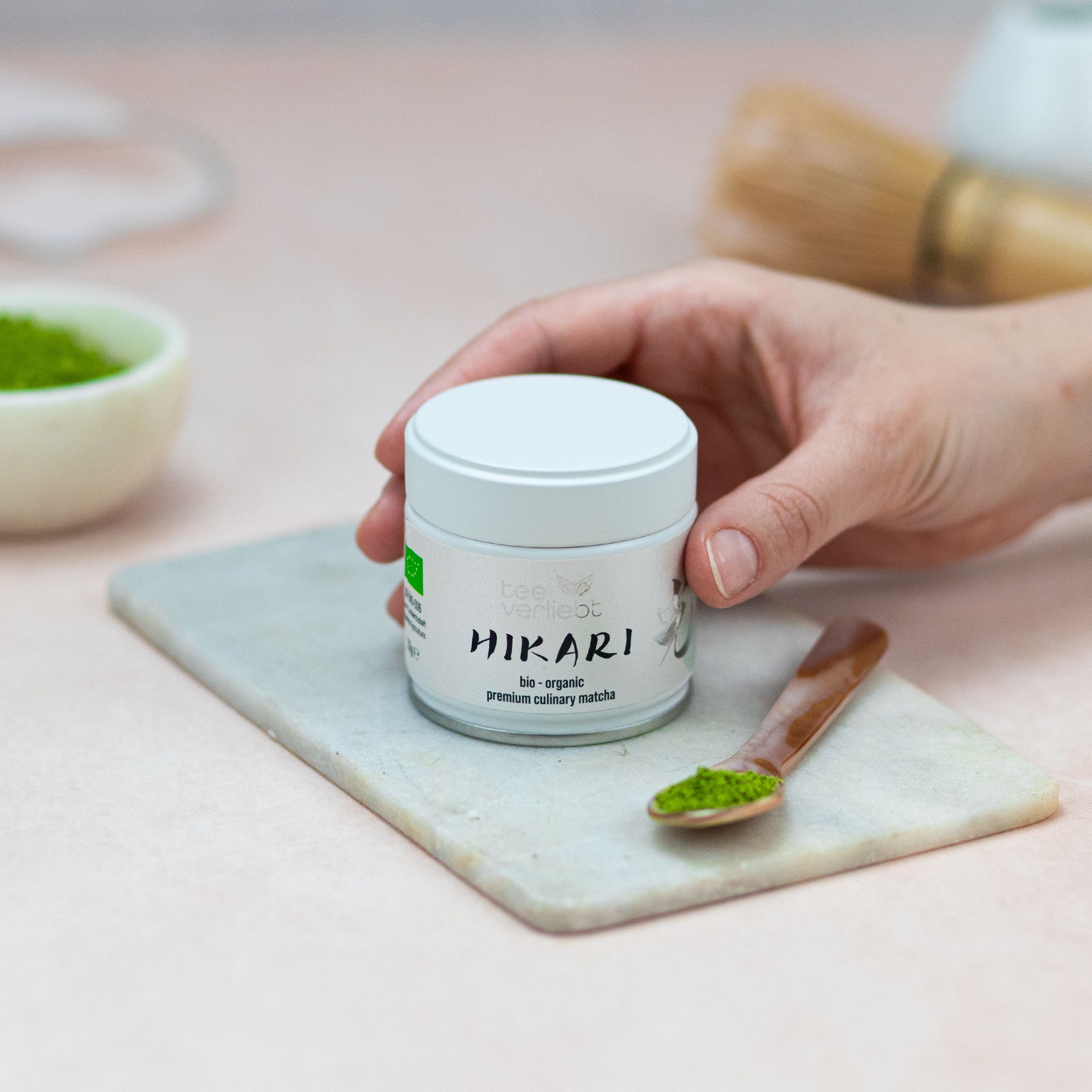 Hand hält Dose "teeverliebt Hikari". Dose auf Marmorplatte. Löffel mit Matcha. Schale mit Matcha-Pulver. Schneebesen im Hintergrund.
