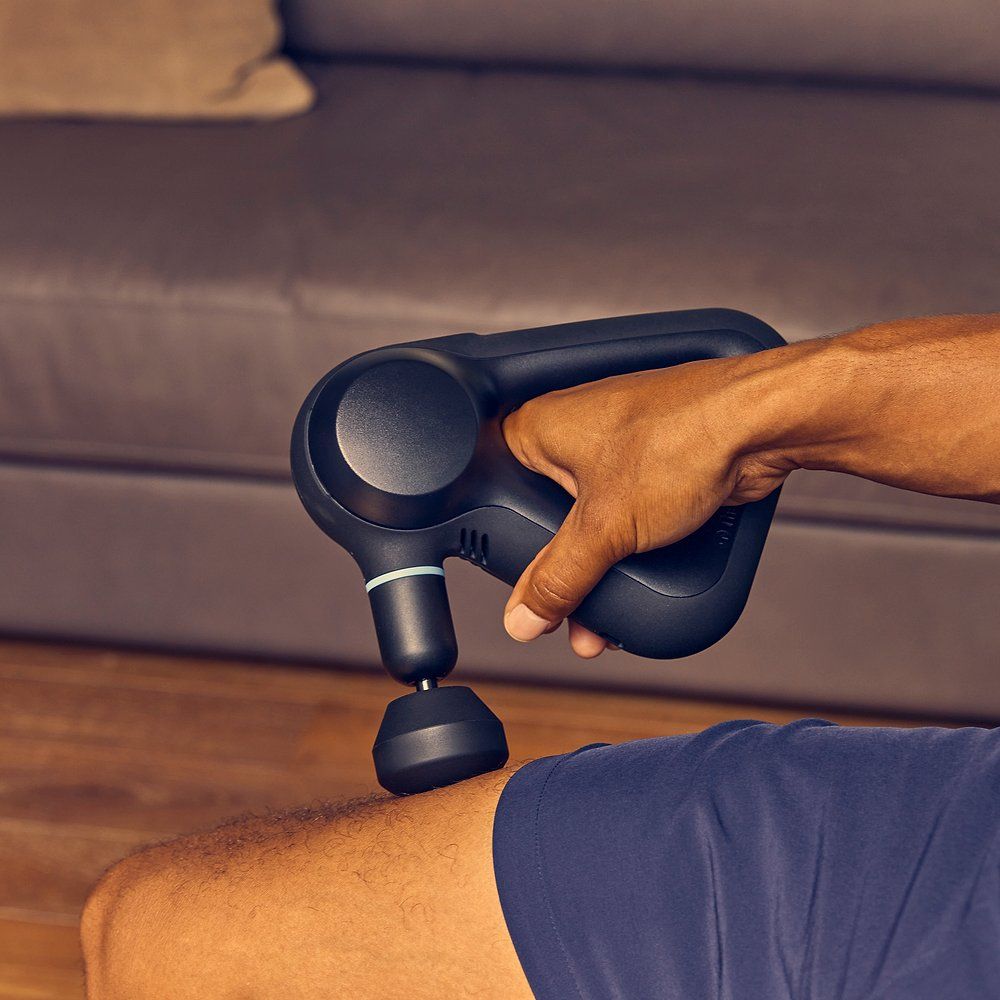 Person hält schwarzes Massagegerät am Bein. Dreieckiger Griff. Aufsatz am Bein. Blaue Akzente.