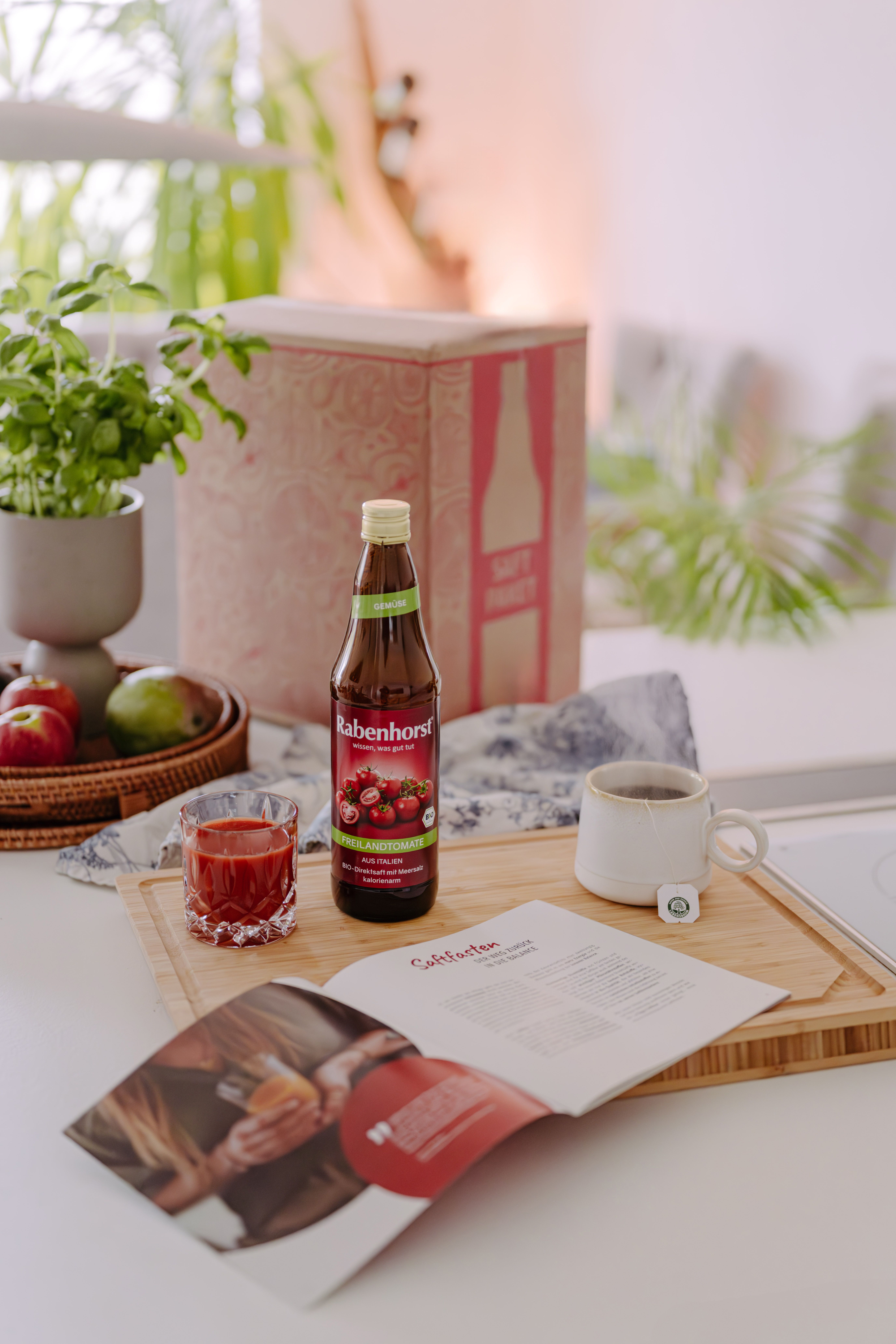 Saftflasche, Glas, Broschüre, Tasse Tee auf Holzbrett. Äpfel und Mango im Korb. Hintergrund: Küche.