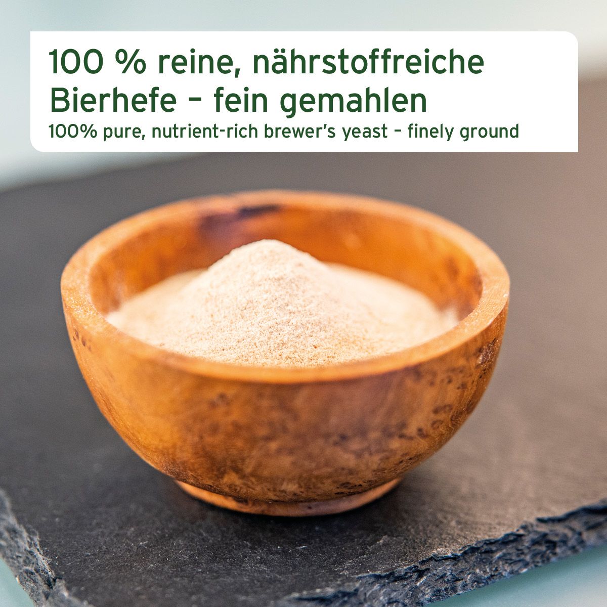Nahaufnahme von fein gemahlener Bierhefe in einer Holzschale. Text: 100% reine, nährstoffreiche Bierhefe.