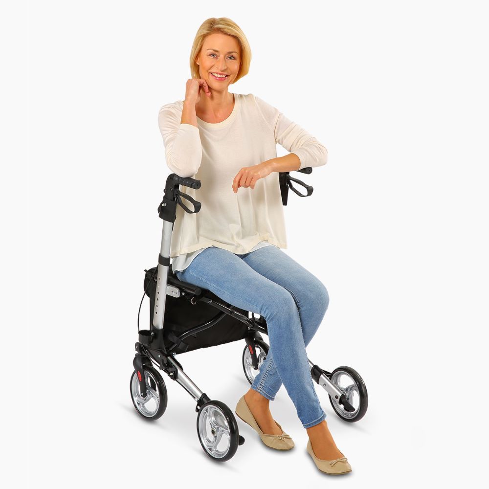 Frau sitzt auf Rollator. Silberfarbenes Gestell, schwarze Sitzfläche und Tasche. Drei Räder.