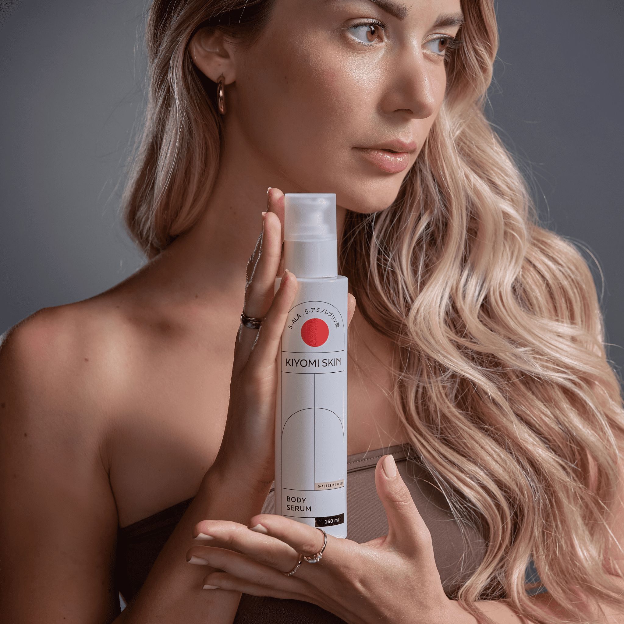 Frau hält eine weiße Flasche mit rotem Kreis und Schriftzug. Produktname: Kiyomi Skin Body Serum. 150 ml.