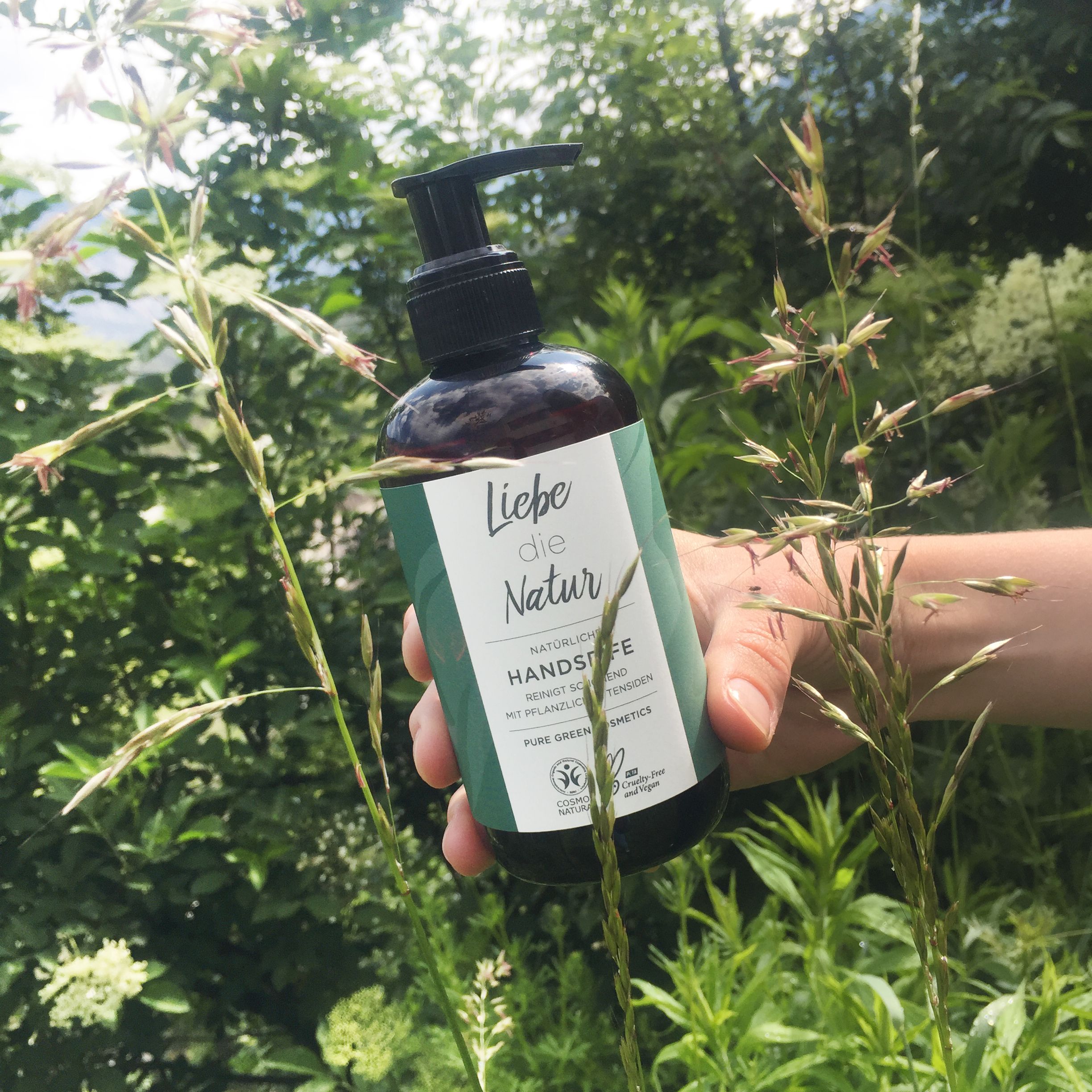Braune Flasche mit schwarzem Pumpkopf. Etikett mit Text: "Liebe die Natur", "Natürliche Handseife". Grün-weißes Design. Hand hält die Flasche.