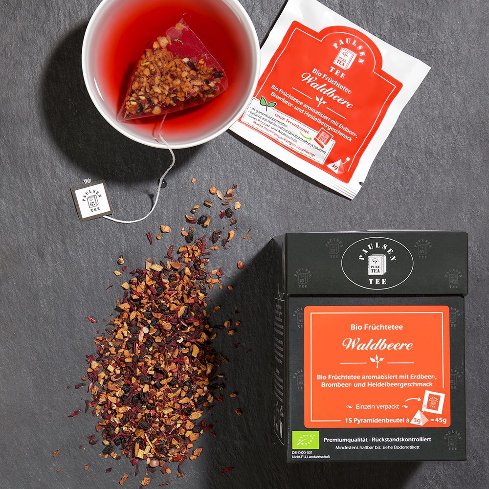 Tasse Tee mit Teebeutel. Daneben: Teebeutel-Verpackung und Box. Aufschrift: Bio Früchtetee Waldbeere. Paulsen Tee Logo. Tee-Blüten.