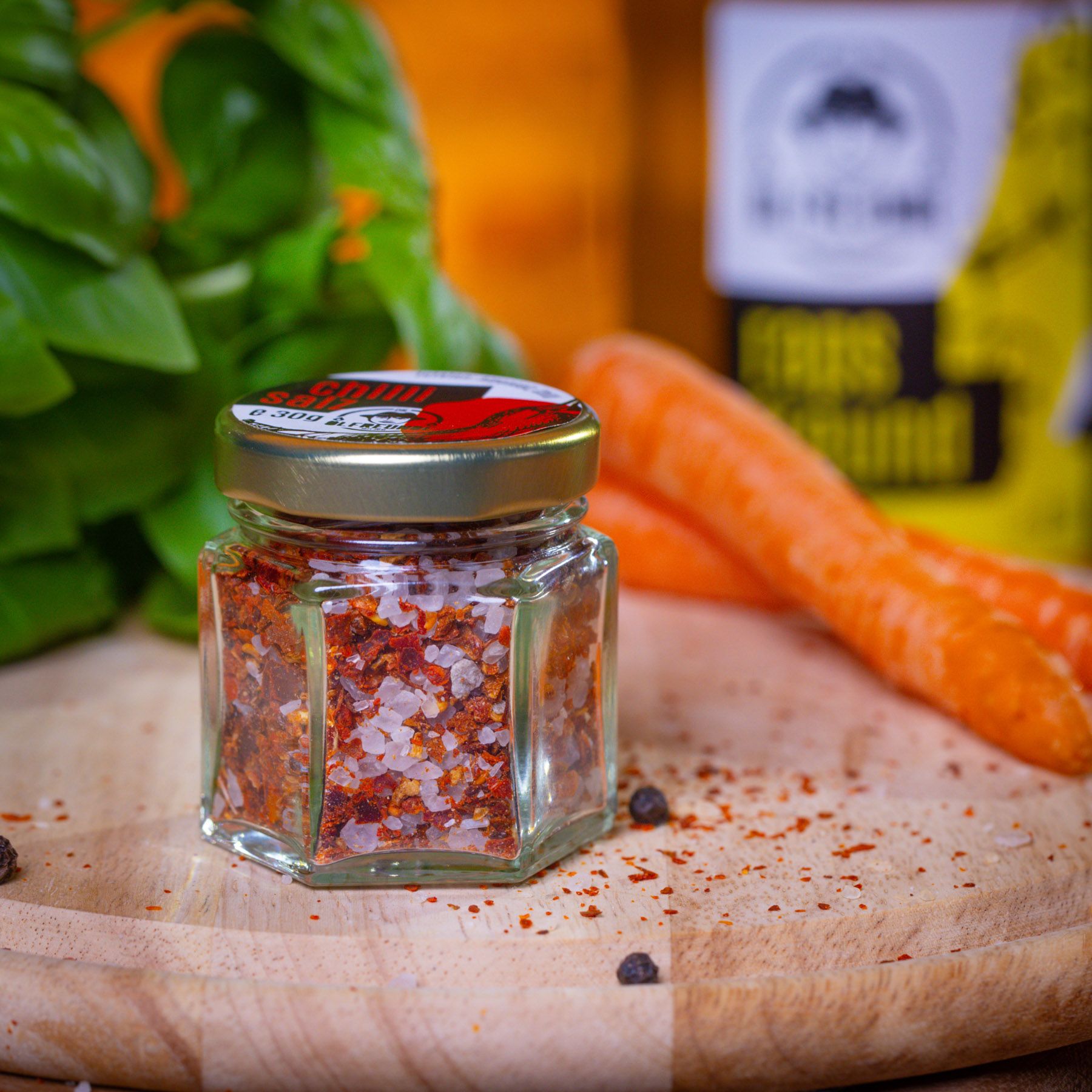 Sechseckiges Glas mit Chilisalz. Deckel mit Chili-Abbildung. Karotten, Basilikum und Pfefferkörner auf Holzbrett. Marke: Die Ölfreunden.