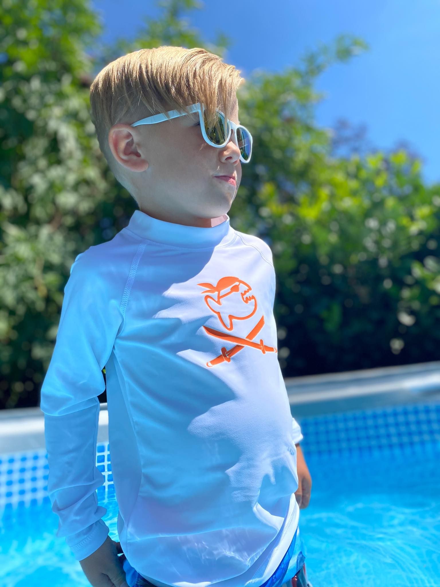 Kind trägt weißes iQ-UV Sonnenschutz Shirt Jolly Kids mit Piraten-Motiv am Pool. Langarm, Stehkragen. Kind trägt Sonnenbrille.