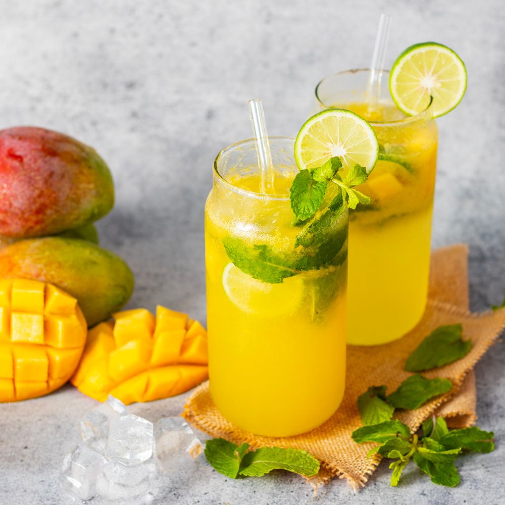 Zwei Gläser mit gelbem Getränk, Limettenscheiben und Minze. Mangos und Strohhalme.