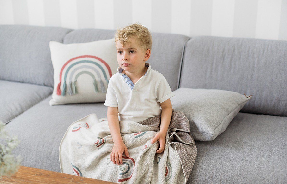 Ein Kind sitzt auf einem Sofa und ist in eine Decke mit Regenbogenmuster gehüllt. Beige mit bunten Regenbogenmotiven.