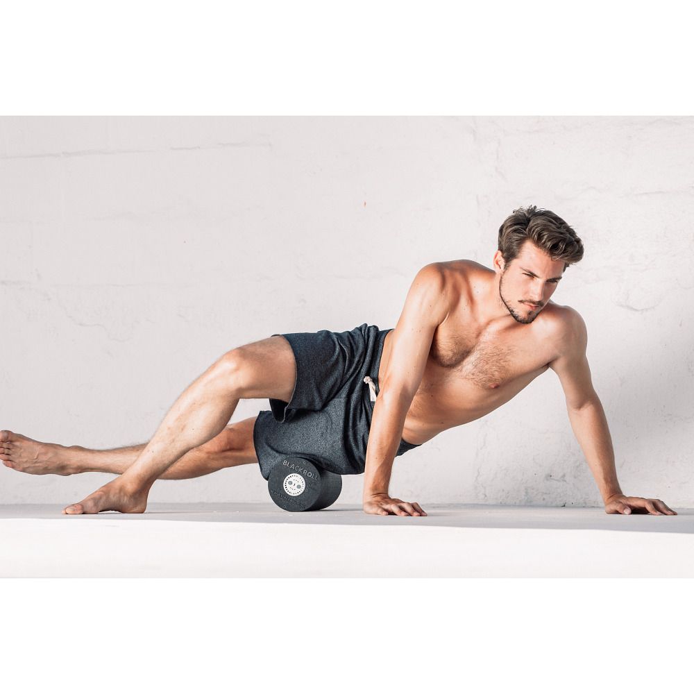 Mann liegt auf einer schwarzen Faszienrolle. Er stützt sich auf einen Arm. Kurze Hose. Helle Umgebung.