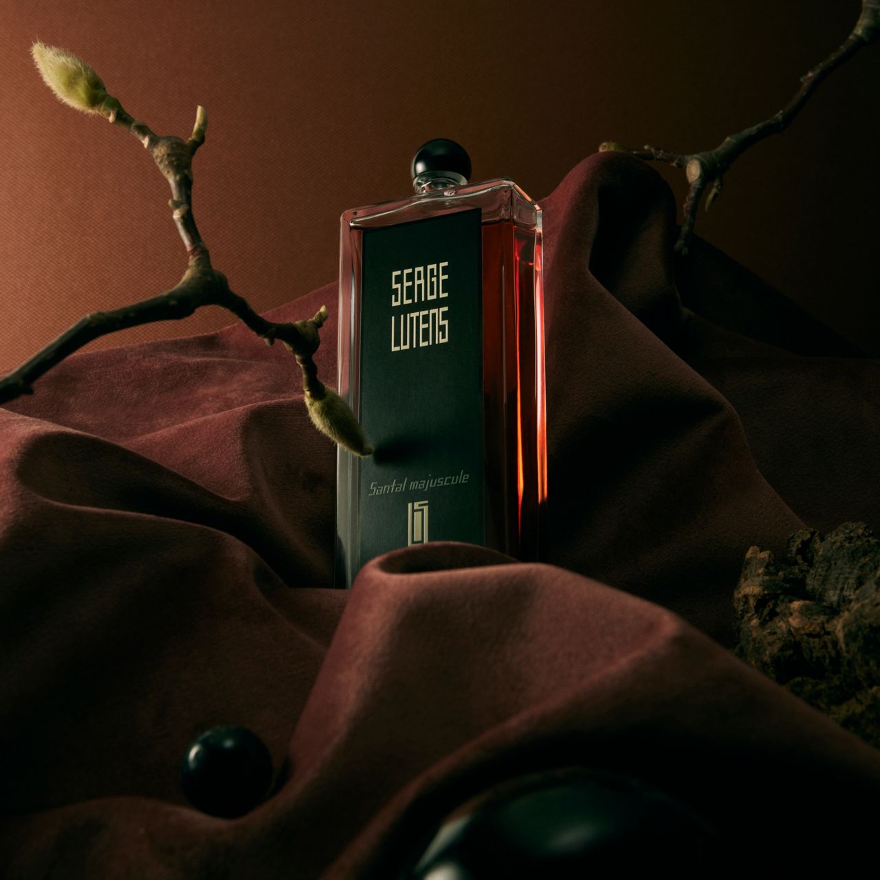 Flakon auf rotem Stoff, mit Ästen. Aufschrift: SERGE LUTENS, Santal Majuscule. Schwarzer Verschluss.