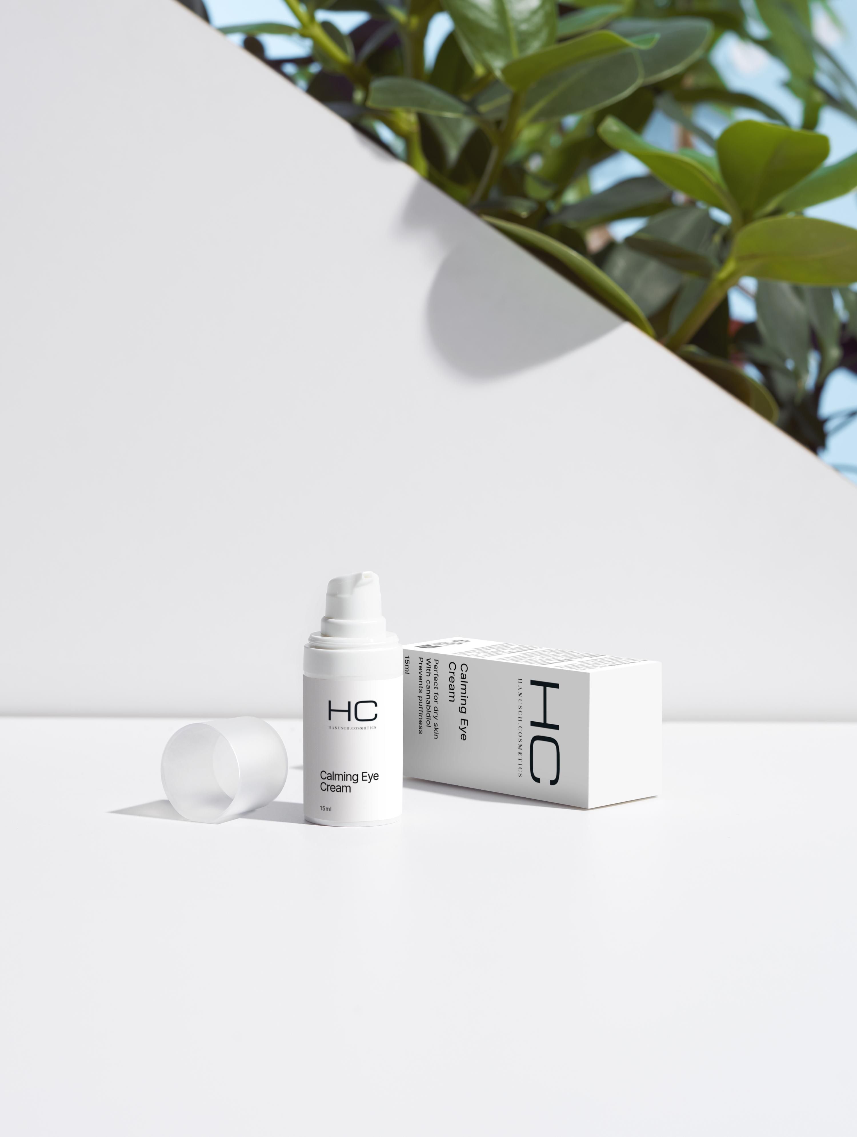 Weiße Flasche "HC Calming Eye Cream" mit geöffnetem Deckel und Karton. Hintergrund: weiße Oberfläche und Pflanzen.