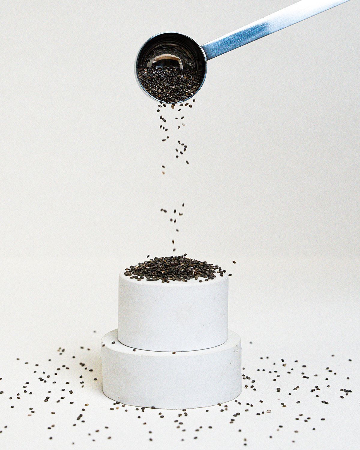 Chia Samen fallen aus einem Löffel auf eine weiße, runde Oberfläche. Mehrere Samen liegen verstreut.