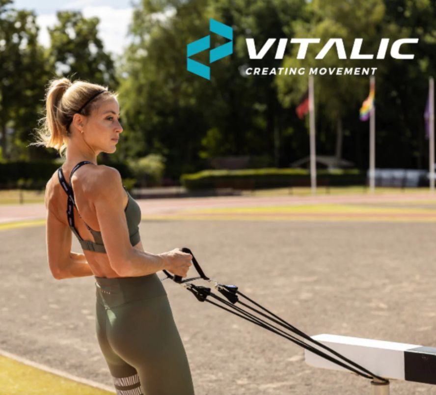 Frau trainiert im Freien mit Widerstandsbändern. Schwarze Bänder mit Griffen. VITALIC-Logo im Hintergrund.
