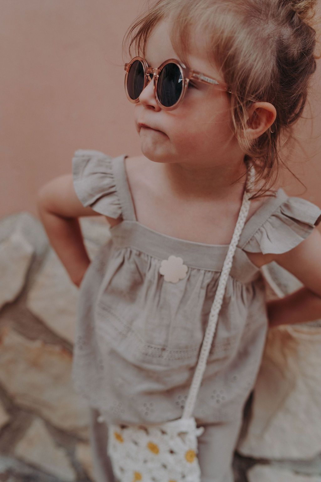 Kleinkind mit Sonnenbrille und Handtasche. Auf dem Oberteil ist ein blütenförmiger Sticker angebracht.
