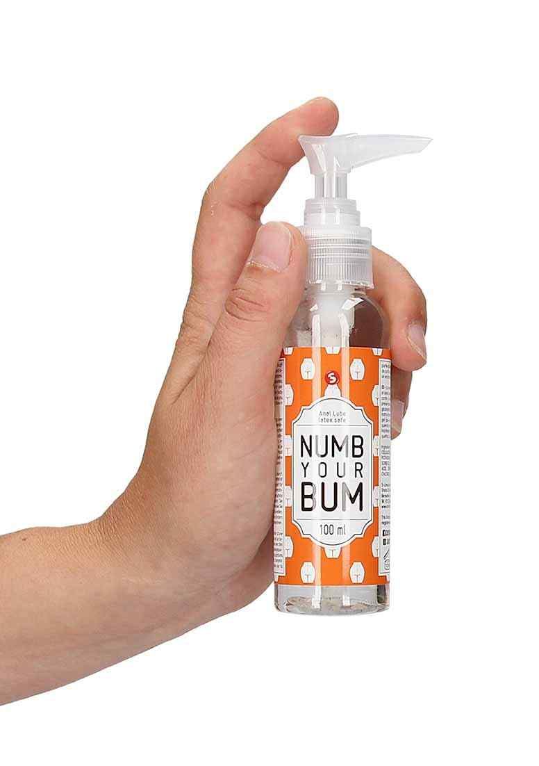 Hand hält transparente Flasche mit Pumpe. Aufschrift: Numb Your Bum. Orangefarbenes Etikett mit weißen Mustern. 100 ml.