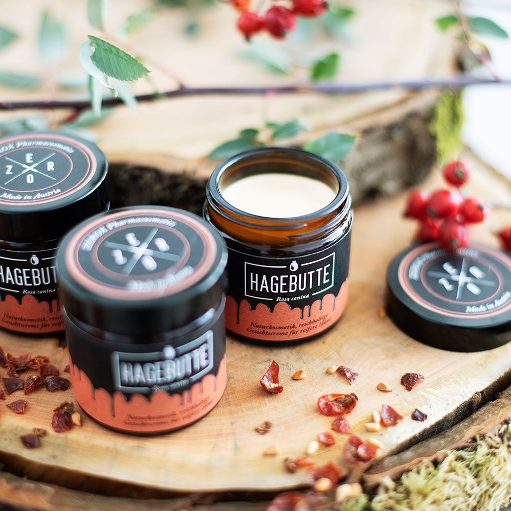 Mehrere Gläser mit schwarzem Deckel und Etikett HAGEBUTTE auf Holz. Geöffnete Dose mit Creme. ZEROX Logo.