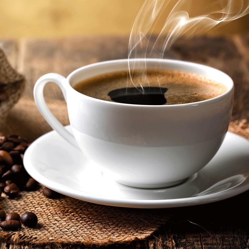 Weiße Kaffeetasse auf Untertasse, gefüllt mit Kaffee. Dampf steigt auf. Kaffeebohnen daneben.