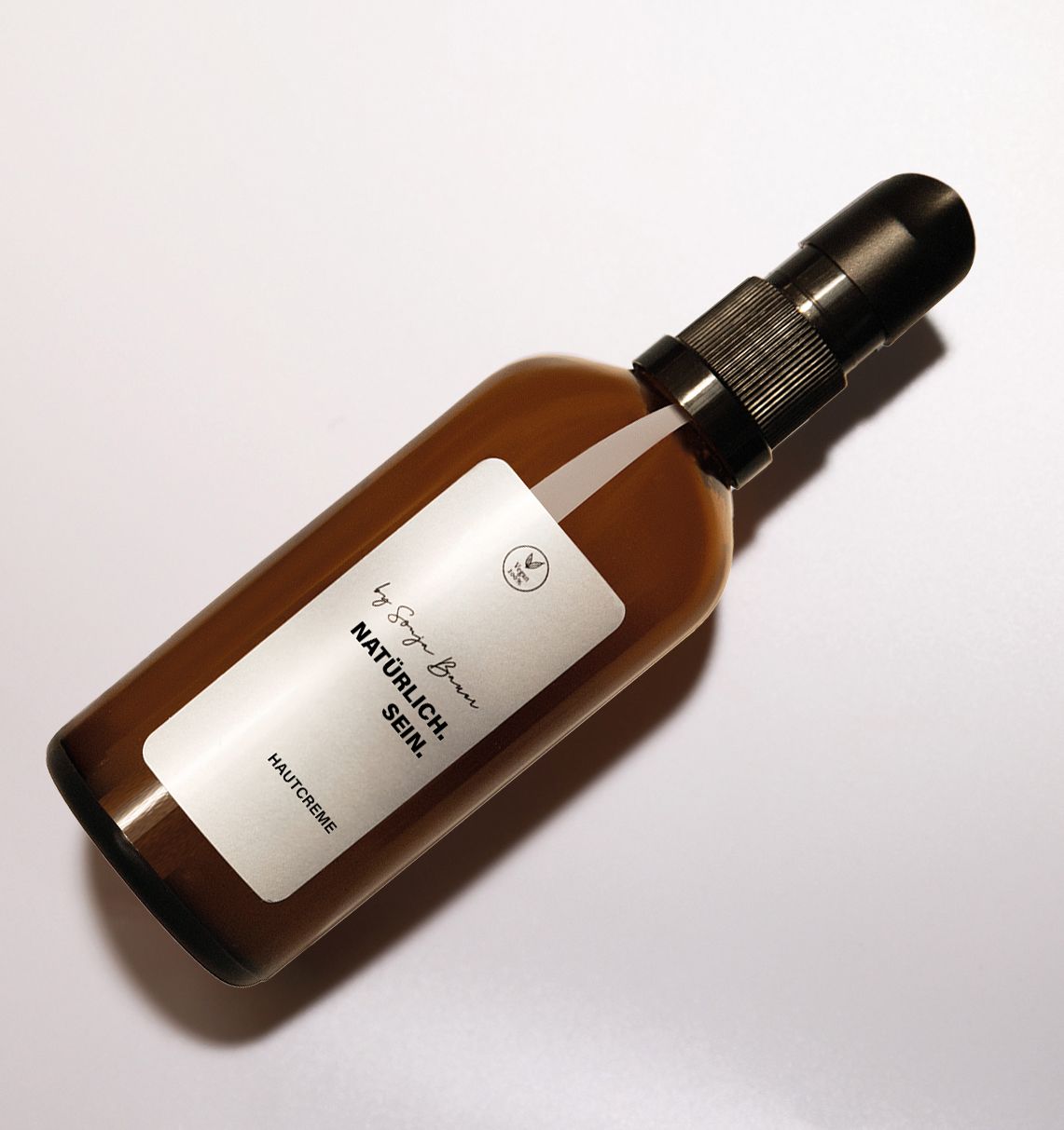 Schräg liegende braune Glasflasche mit schwarzem Pumpkopf. Weißes Etikett mit Text: NATÜRLICH.SEIN. und HAUTCREME. Marke: By Sonja Bauer.