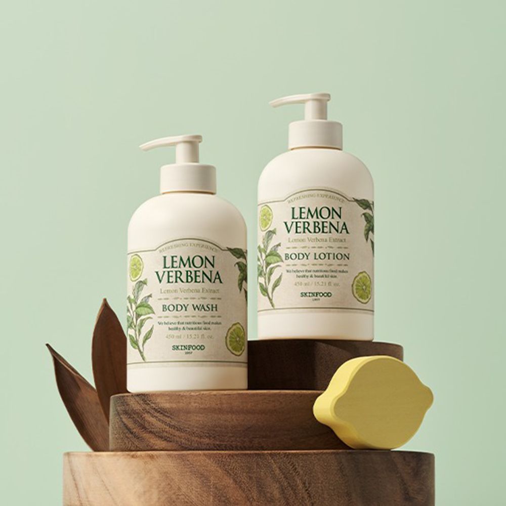 Zwei "Lemon Verbena" Produkte: Körperwaschgel und Body Lotion. Beige Flaschen mit Pumpverschlüssen. Auf Holzpodest, mit Deko.