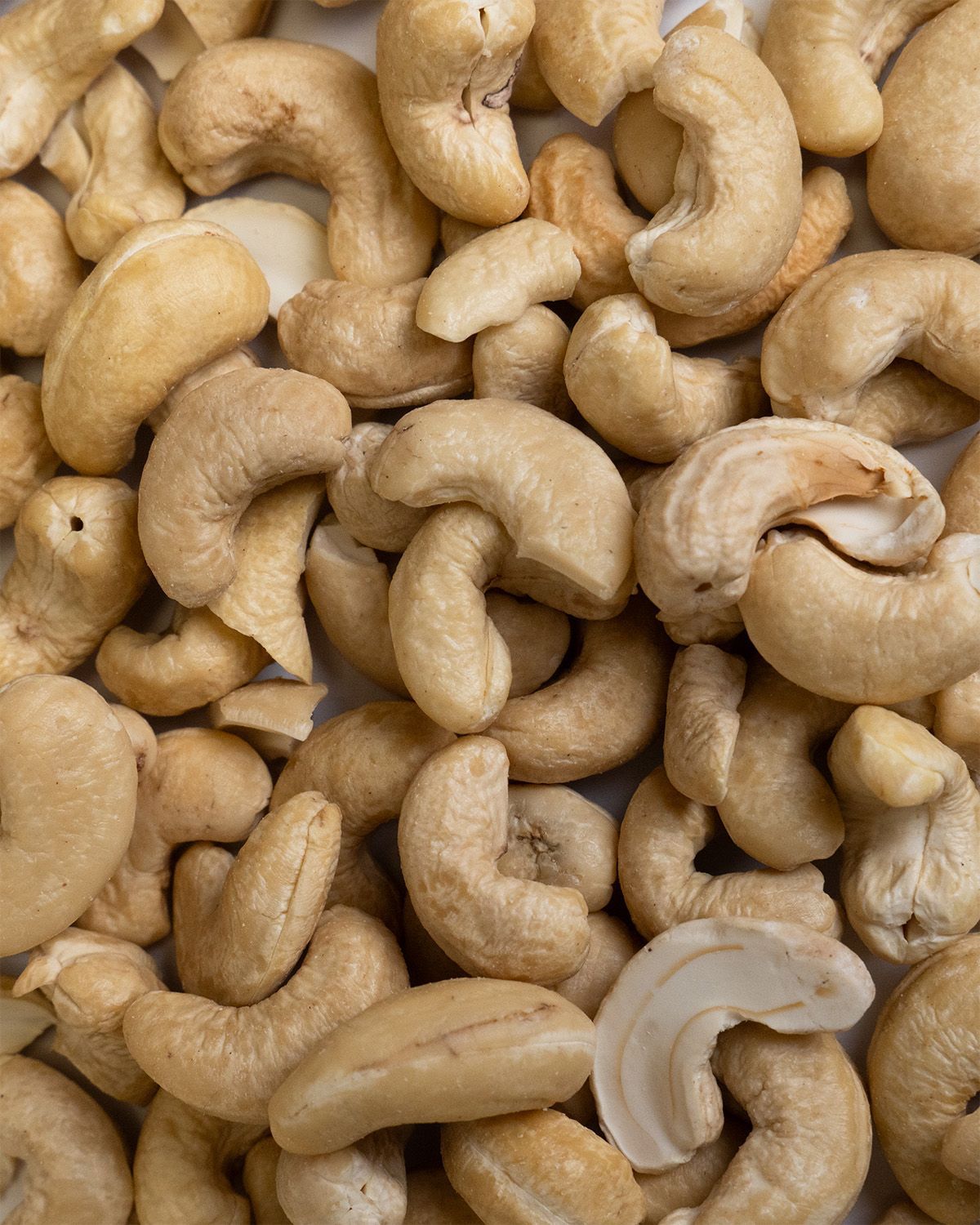 Nahaufnahme von Cashewkernen. Helle, beige Nüsse, ungeordnet angeordnet. Einzelne Kerne sind angeschnitten.