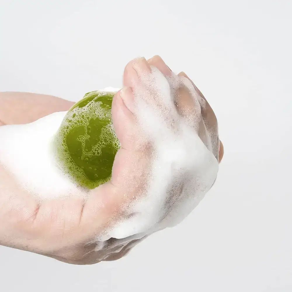 Grünliche Kugel in Hand, umgeben von Schaum. Helle, runde Form. Hände im Fokus.