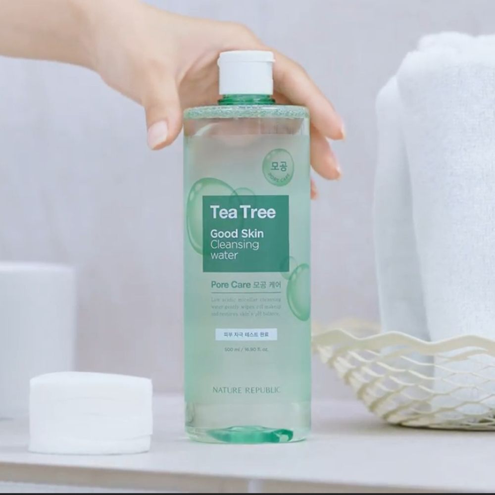 Produktflasche mit grünem Etikett. Text: Tea Tree Good Skin Cleansing Water. Hand hält Flasche. Wattepads und Handtuch im Hintergrund.