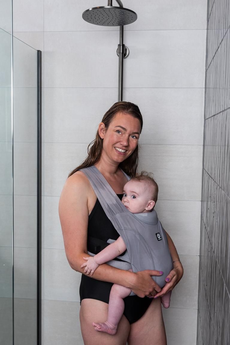 Frau mit Baby in grauer Babytrage unter der Dusche. Dooky-Logo sichtbar.