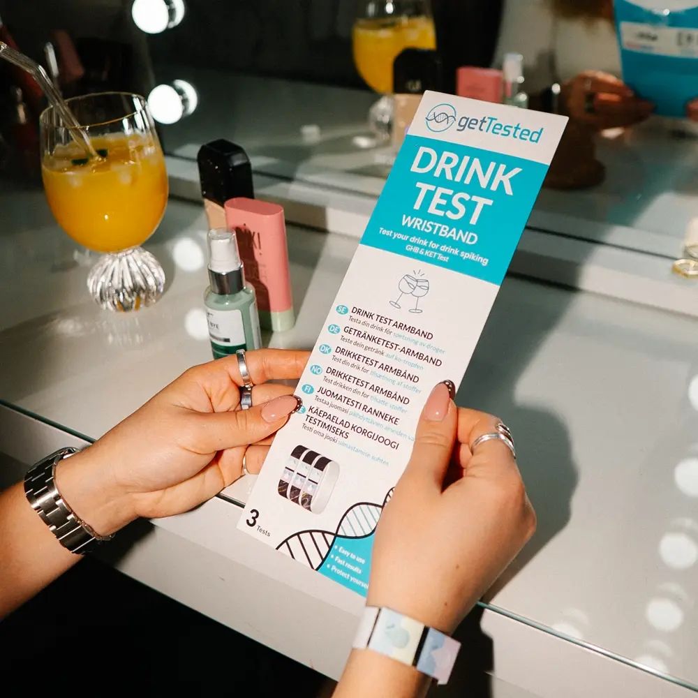 Person hält eine Verpackung mit "GetTested Drink Test Wristband". Mehrere Armbänder sind sichtbar. Hintergrund: Schminktisch.