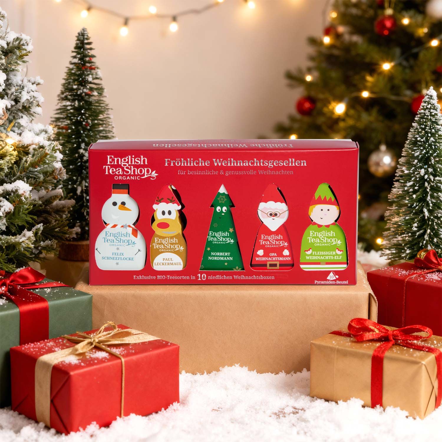 Rote Geschenkbox mit 5 weihnachtlichen Tee-Anhängern, umgeben von Geschenken und Weihnachtsbäumen.
