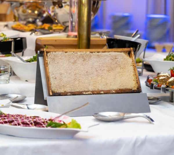 Honigwabe in Holzrahmen auf einem Buffet. Die Wabe ist auf einem Ständer platziert, umgeben von Speisen und Utensilien.