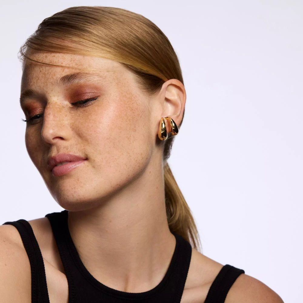 Porträt einer Frau mit geschminkten Augen. Warme Farbtöne. Goldene Creolen. Blonde Haare, Pferdeschwanz.