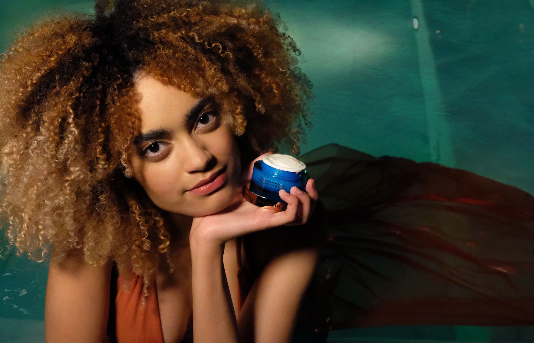 Frau hält einen Tiegel in der Hand. Produktname und Text sichtbar. Creme im Inneren.