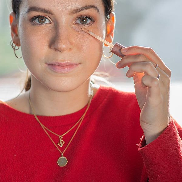 Frau trägt GRN Liquid Concealer beige oat auf. Roter Pullover, goldene Halsketten. Applikator wird unter dem Auge verwendet.