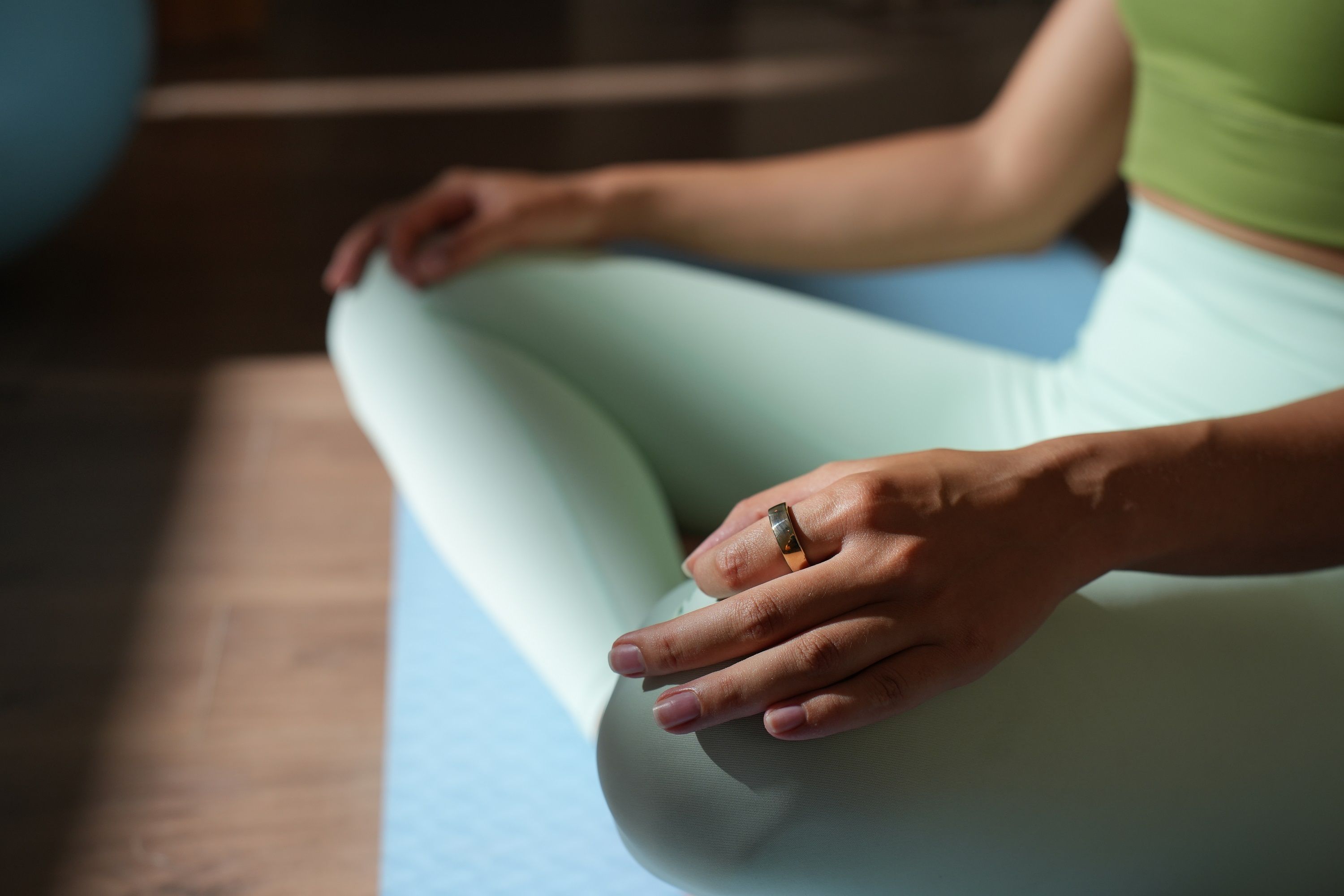 Person sitzt auf einer Yogamatte. Sie trägt einen Ring am Finger. Helle Kleidung.