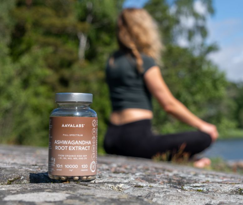 Aavalabs Ashwagandha Flasche auf Felsen, Frau meditiert im Hintergrund.
