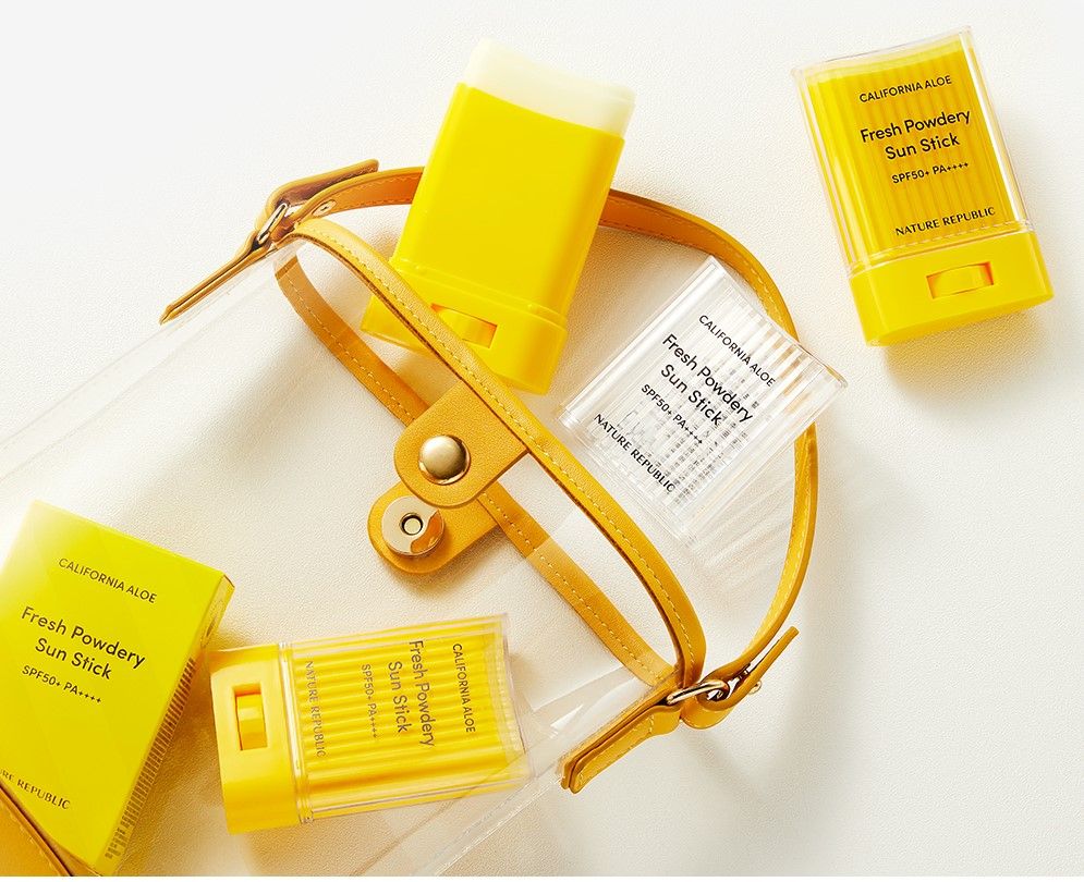 Mehrere gelbe Sonnensticks und Verpackungen in einer transparenten Tasche mit gelbem Griff. Text: Fresh Powdery Sun Stick.