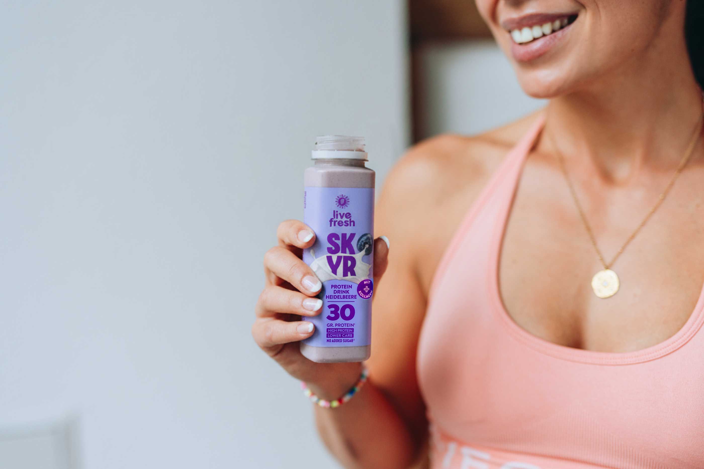 Frau hält eine Flasche Skyr Drink. Auf der Flasche: Logo, „SKYR“, „30 gr. Protein“, „Heidelbeere“.