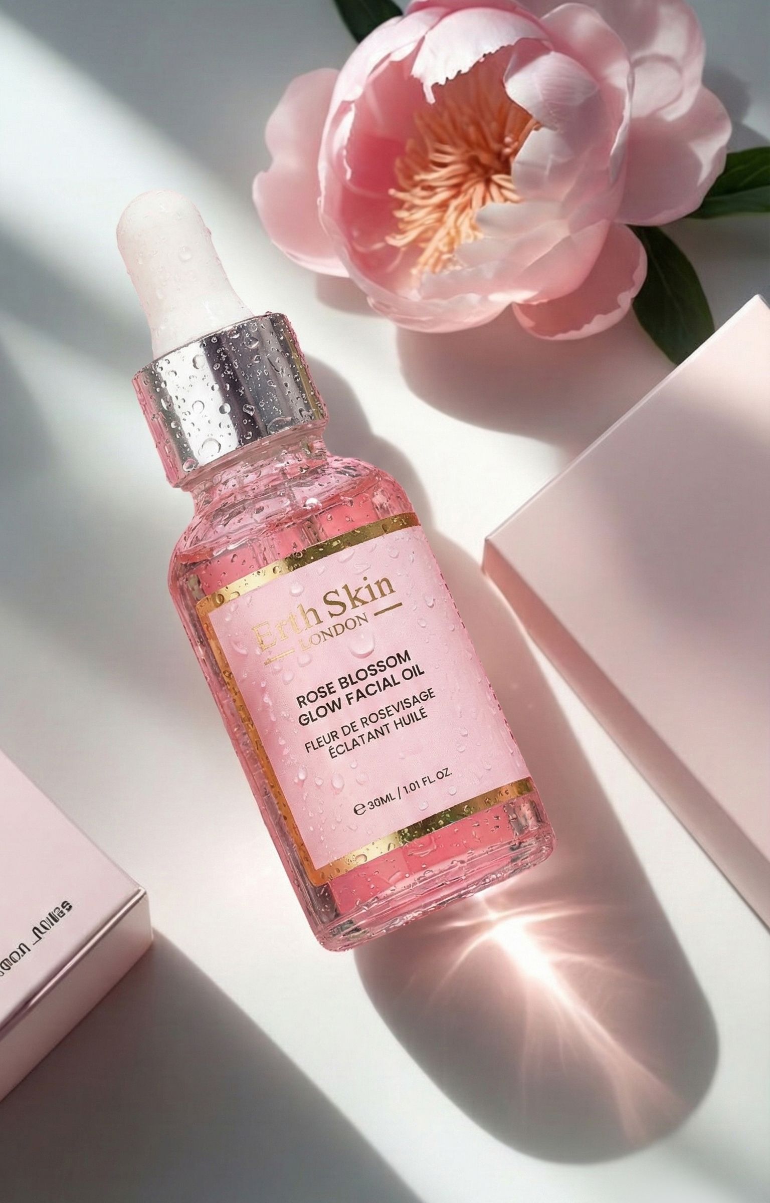 Glasflasche mit rosafarbenem Gesichtsöl, Tropfen auf der Oberfläche. Rosa Box und Blüten im Hintergrund.