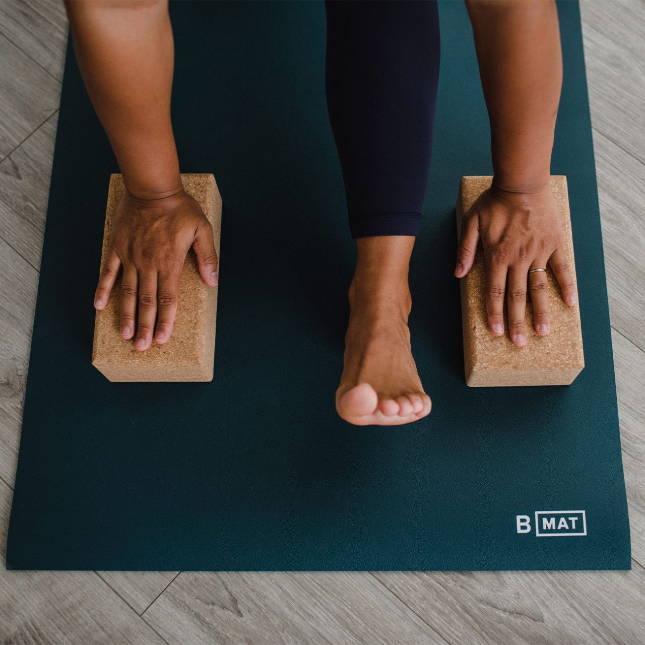 Person in Yoga-Position auf dunkelgrüner Yogamatte. Hände auf Holzblöcken. "B MAT"-Logo sichtbar.