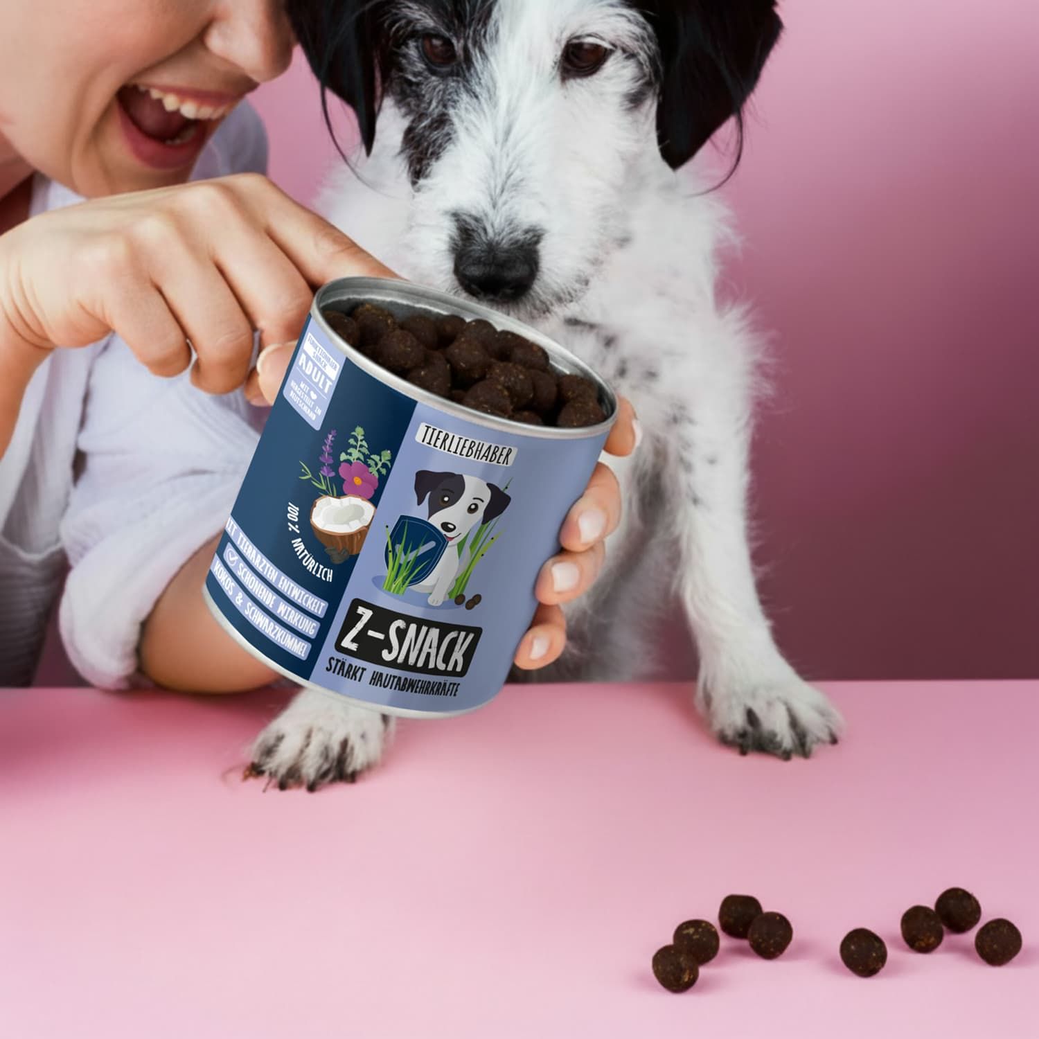 Person hält Dose mit Z-Snack-Kugeln. Hund schaut zu. Dose mit Hund-Illustration. Rosa Hintergrund.