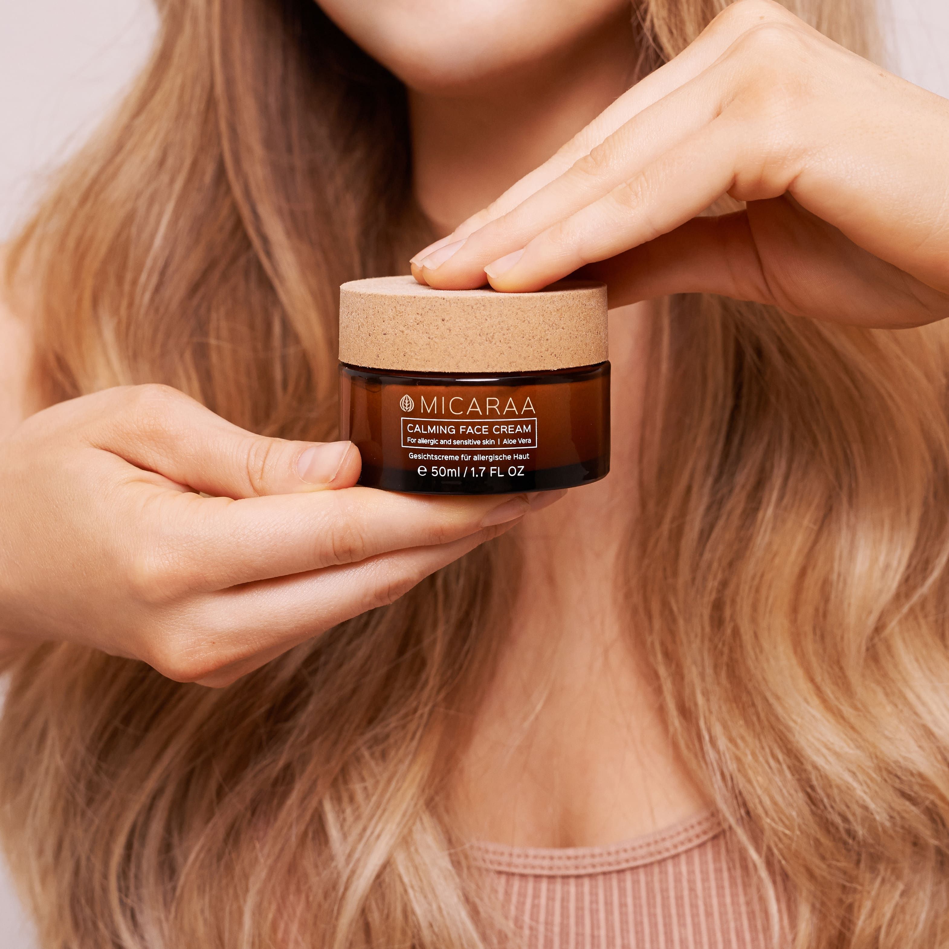 Person hält eine braune Creme-Dose mit Holzdeckel. Aufschrift: MICARAA Calming Face Cream. Für allergische Haut. Mit Aloe Vera. 50ml.