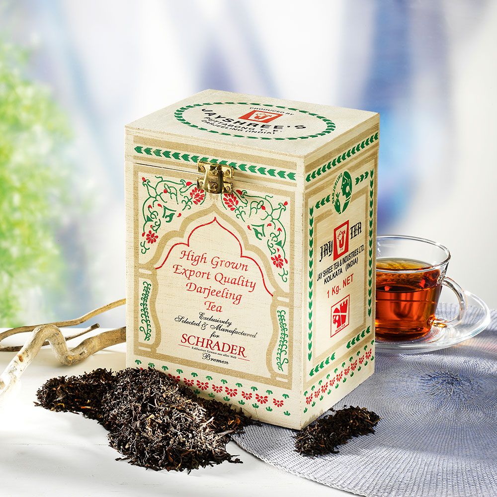 Holzkiste mit Aufschrift 'Schrader Darjeeling Tee'. Daneben eine Tasse Tee und lose Teeblätter.