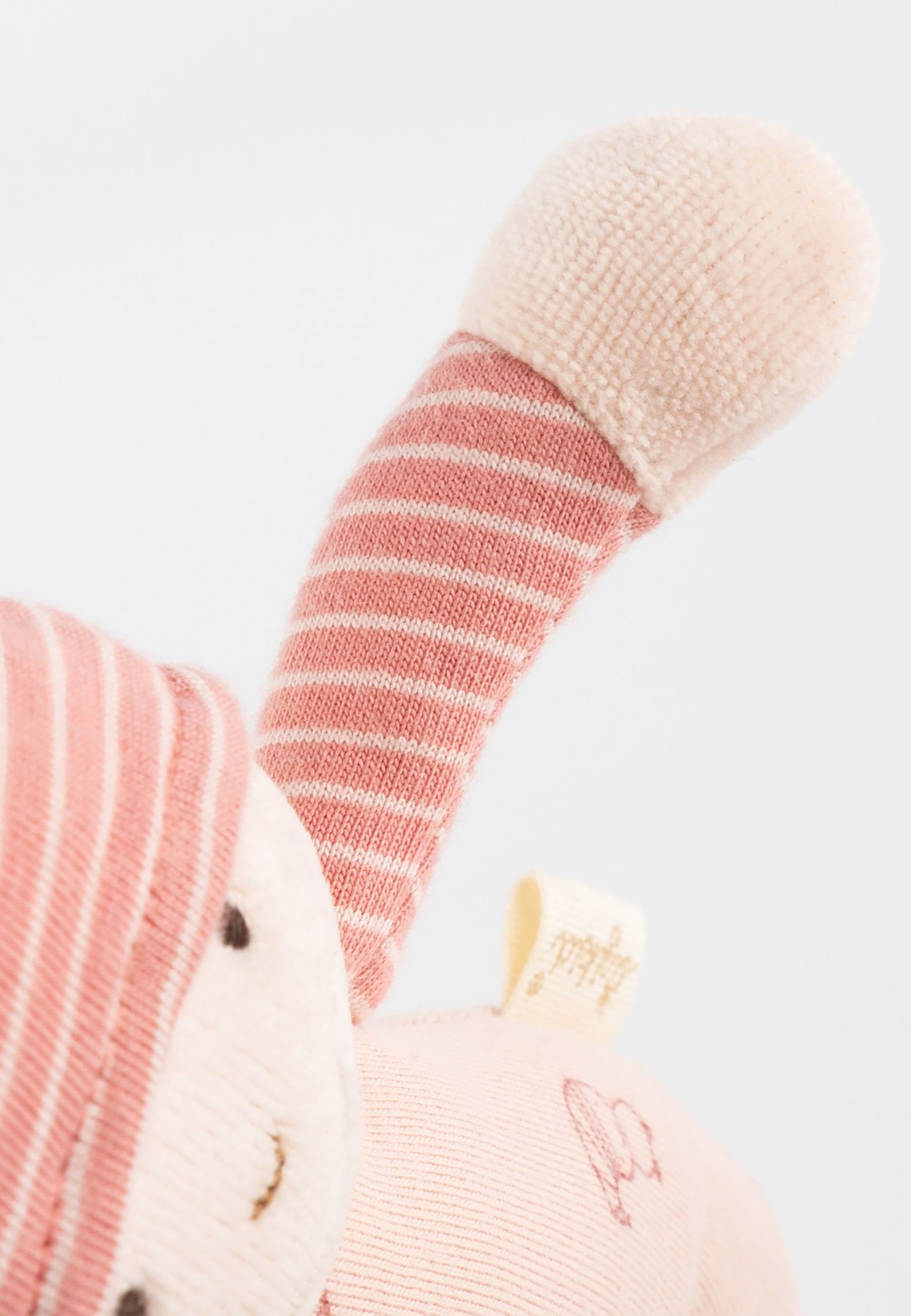Nahaufnahme der Puppe. Rosa Stoff, gestreifte Arme und Mütze. Aufgesticktes Gesicht. Etikett mit Logo.