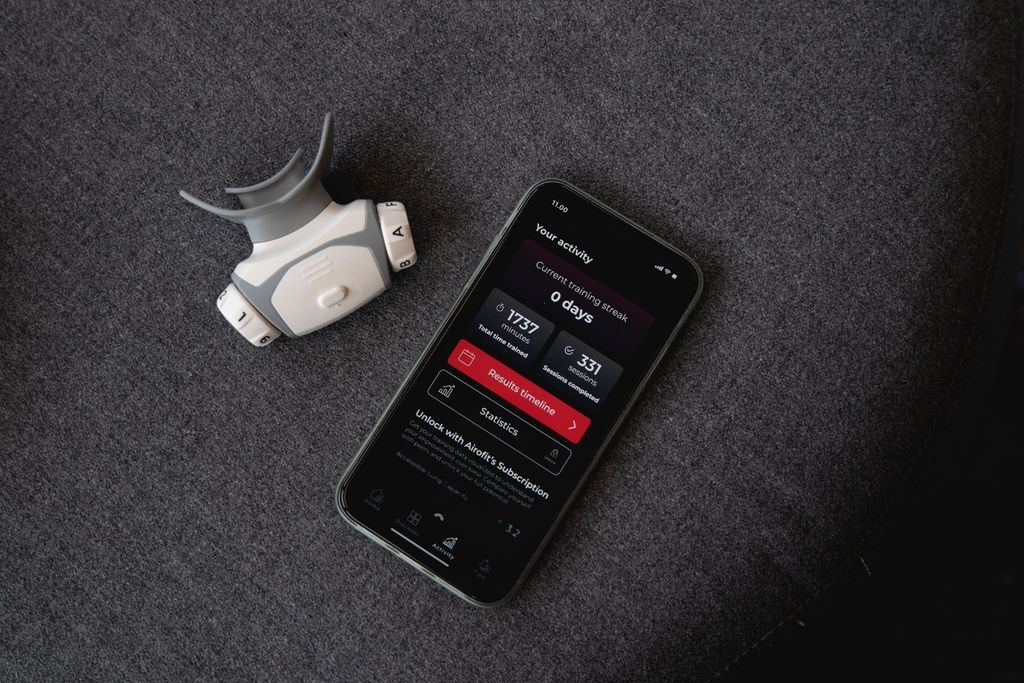 Atemtrainer und Smartphone auf grauer Oberfläche. Smartphone zeigt App-Oberfläche mit Trainingsdaten. Airofit-Logo sichtbar.