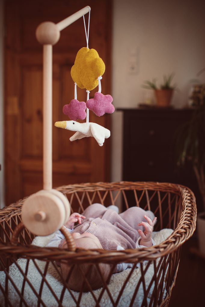 Mobile über Baby in Weidenkorb. Gelbe Wolke, rosa Wolken, weiße Gans. Holzarm und -fuß.