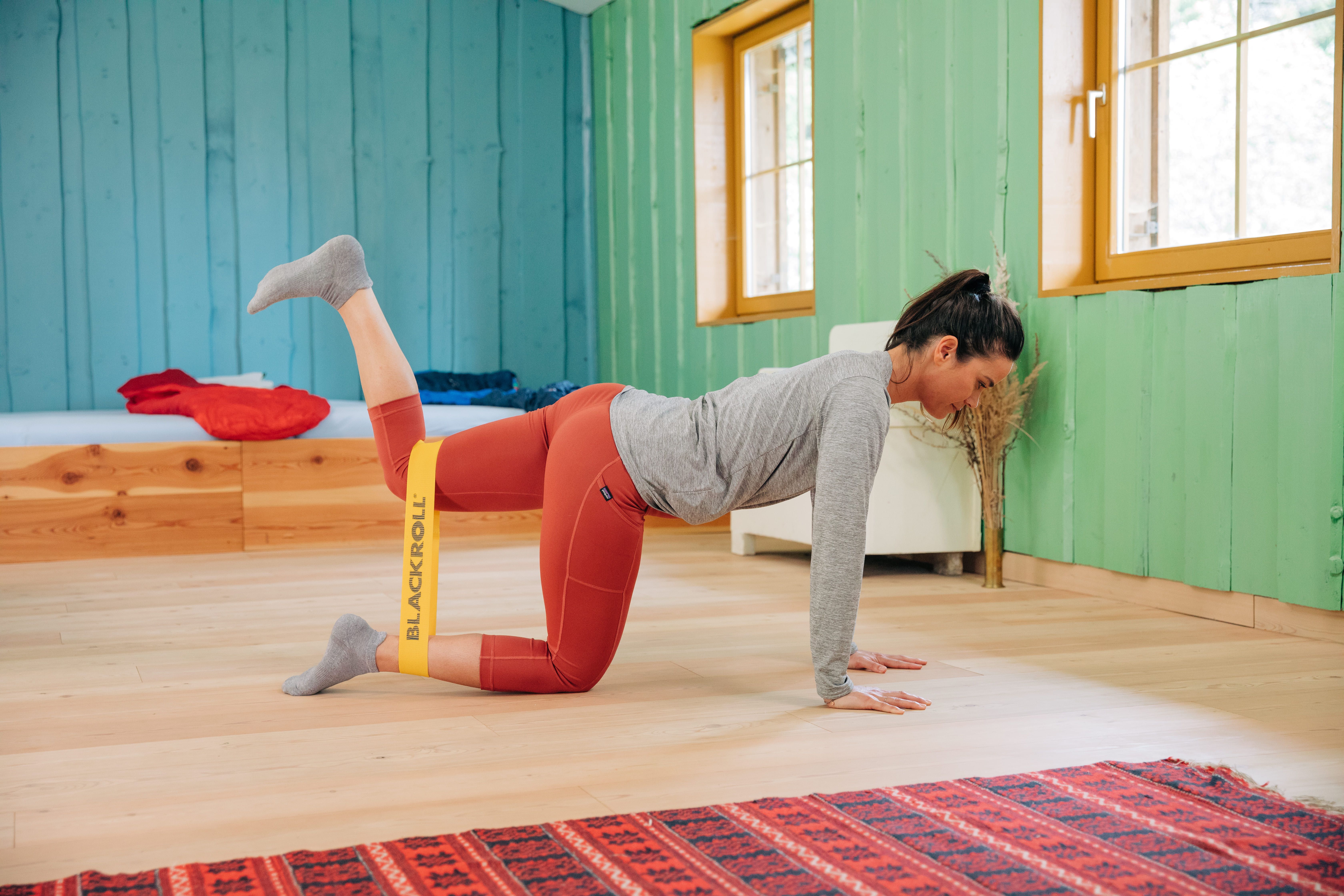 Frau in Fitness-Pose mit gelbem BLACKROLL Loop Band um die Beine. Rote Hose, graues Oberteil.