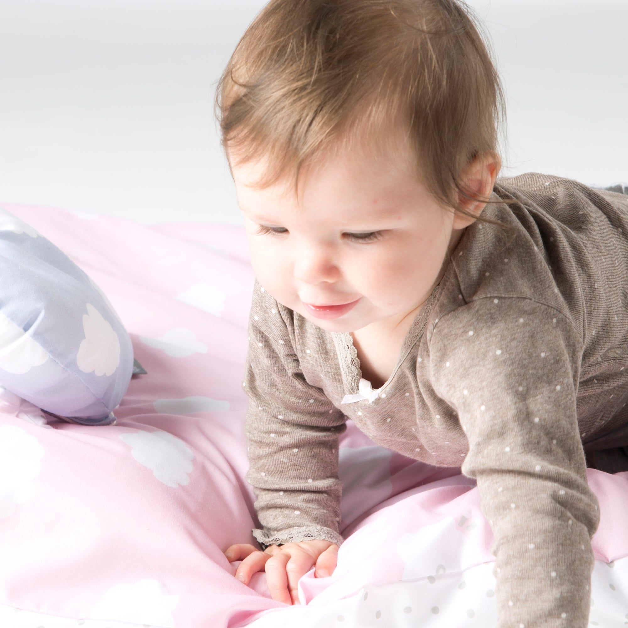 Baby krabbelt auf rosa Bettwäsche mit Wolkenmuster. Baby trägt braunen Strampler.