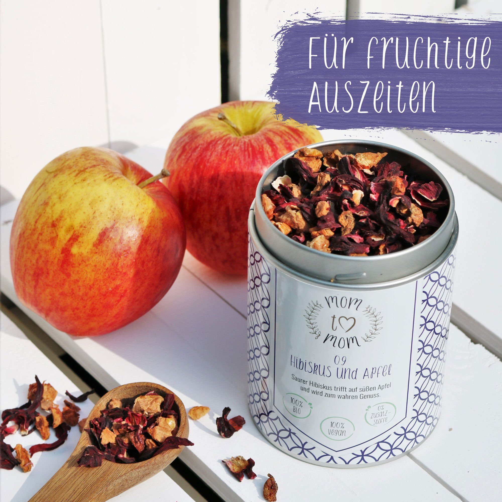 Teedose mit losem Tee, zwei Äpfel und Holzlöffel mit Tee. Dose mit Aufschrift 'mom to mom' und 'Hibiskus & Apfel'.